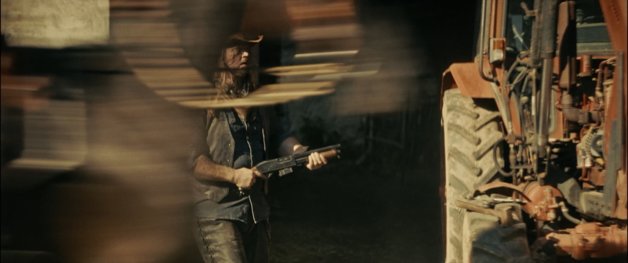 Long-haired man in a denim vest gripping a pump-action shotgun beside an orange tractor, motion blur - Amon Amarth